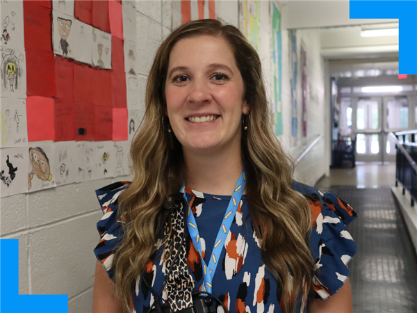 Courtney Ellis stands in a hallway in front of student artwork, smiles at the camera.